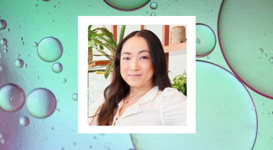 A woman with long dark hair and a warm smile, wearing a white blouse, poses for in front of indoor green plants. The image is contained in a square white frame. The background features an abstract design with turquoise water droplets and pinkish bubbles.