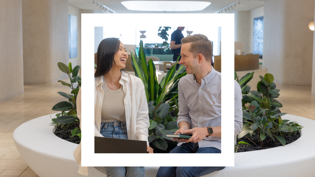 A Polaroid-style photograph features two people seated next to each other in a modern, indoor space with neutral tones and contemporary decor. They are sitting on the edge of a circular planter filled with lush green plants. The woman on the left, wearing a white shirt and jeans, is laughing while looking at the man. The man on the right, wearing a light blue striped shirt, is smiling back at her while holding a tablet. The background shows another person standing and working in the spacious, well-lit room, adding to the professional and friendly atmosphere.